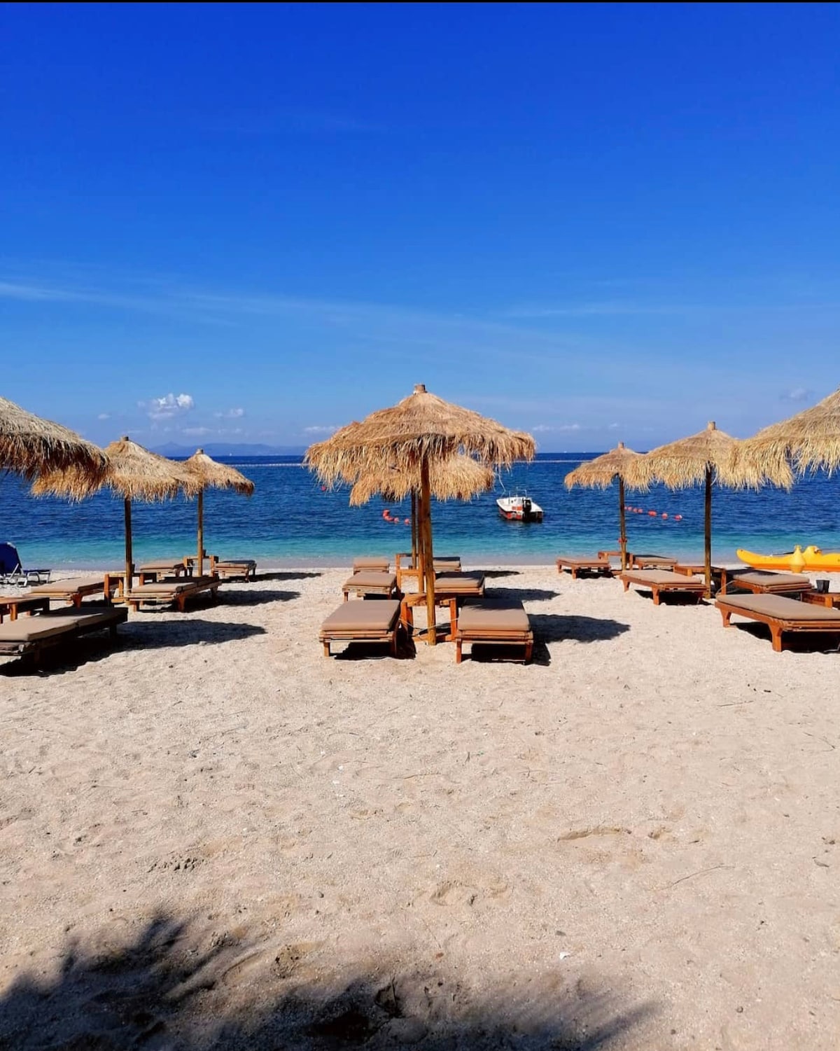 A sandy beach is lined with wooden loungers and straw umbrellas, creating a relaxing atmosphere. Clear blue waters stretch into the distance, punctuated by a few boats in the waves. The bright sky enhances the beach's inviting appearance.