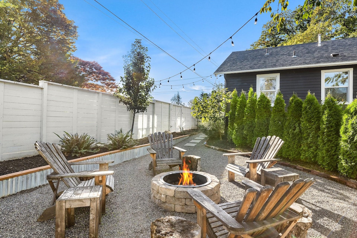 A private backyard features a circular fire pit surrounded by four wooden adirondack chairs. The space is lined with gravel and bordered by a white fence, with greenery and trees in the background, while string lights create an inviting ambiance overhead.