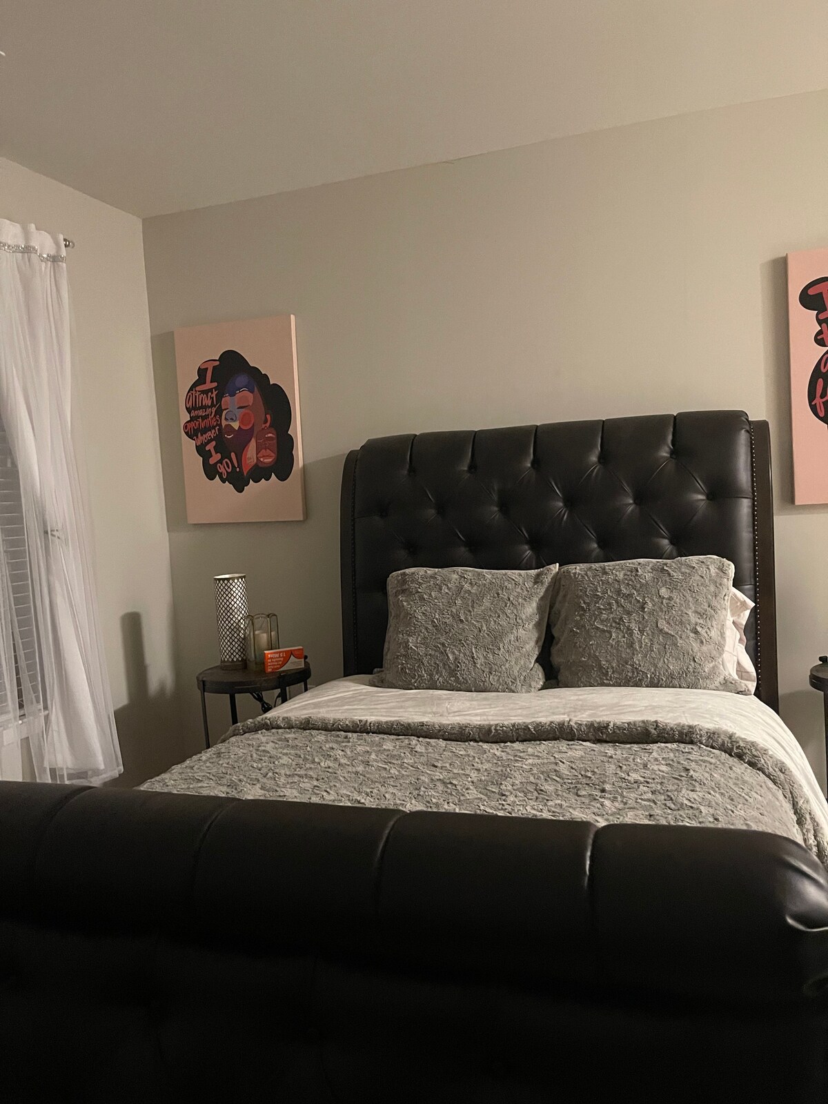 A comfortable sleeping area features a padded, dark headboard with plush pillows. Light-colored bedding is neatly arranged on the bed. On either side, accent tables hold decorative items, while two colorful wall art pieces add a creative touch to the space.