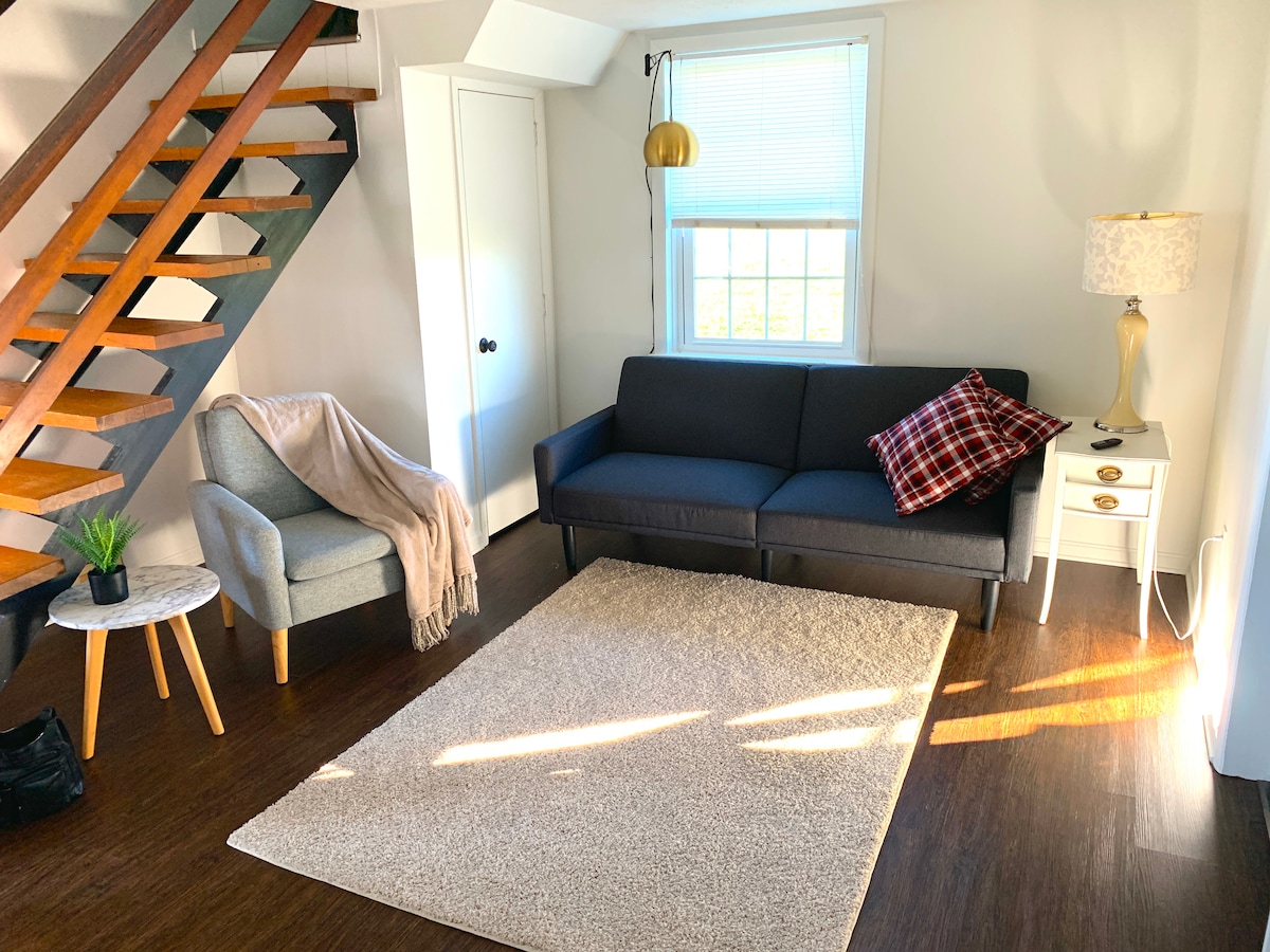 A cozy living area features a dark sofa with a plaid throw pillow and a stylish lamp beside it. A light rug covers the floor, while an armchair with a soft blanket and a small plant add warmth. Natural light fills the space through a window.