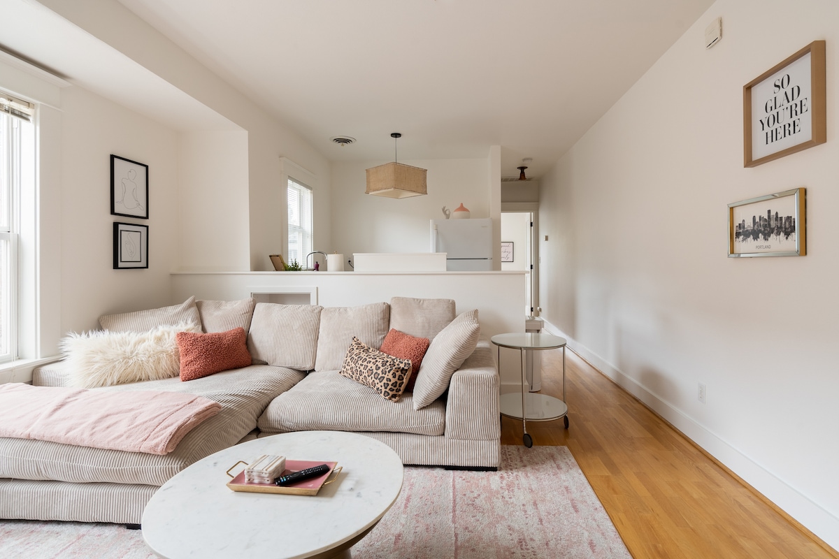 A spacious living area features a light-colored sectional sofa adorned with decorative pillows and a cozy throw. A marble coffee table sits in front, enhancing the modern aesthetic. The bright, neutral walls and abundant natural light create an inviting atmosphere, with minimalist artwork displayed on the walls.