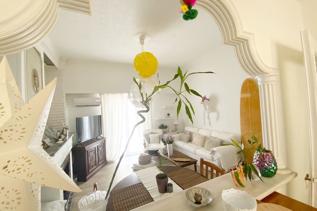 A bright living area is visible, featuring a large cream sofa accented with neutral pillows. Natural light floods the space through sheer curtains, while a sculptured fireplace adds character. Delicate decorative elements, including a star-shaped ornament and potted plants, enhance the inviting ambiance.