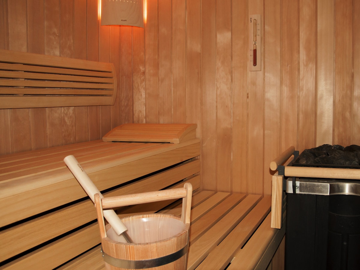 An interior view of a wooden sauna is presented, featuring benches arranged along the walls. A thermometer and a wooden bucket sit nearby, indicating the sauna's essential amenities. Warm wood tones dominate the space, creating a calming environment for relaxation.