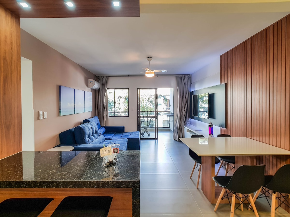 An open living area is presented, featuring a spacious blue sofa positioned against a wall. A large window allows natural light, with access to a balcony visible. A dining table with black chairs sits adjacent to a modern kitchen counter, creating a contemporary space.