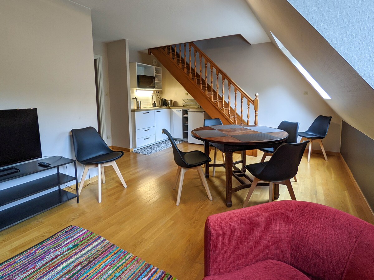 A bright and airy living area features a red sofa and a circular wooden dining table surrounded by four black chairs. Modern kitchen amenities are visible in the background, with stairs leading to an upper level. Natural light floods the space through slanted ceilings.