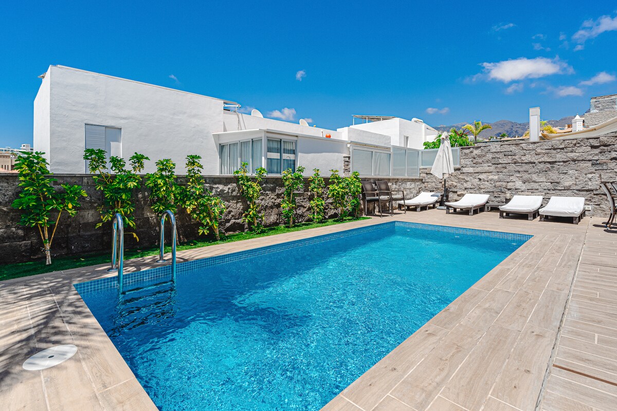 A private heated swimming pool is surrounded by a wooden deck and lush greenery. Sun loungers are positioned for relaxation, while a shaded area is provided by an umbrella. Clear blue skies are visible above, enhancing the space's inviting atmosphere.