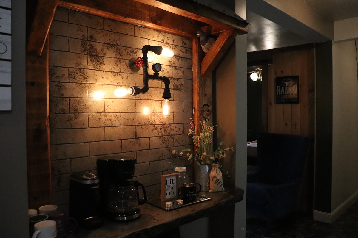 A rustic coffee station features vintage-style lighting above a wooden shelf. A coffee maker and a kettle rest on the counter, accompanied by various mugs. Decorative elements, including a vase of dried flowers, are arranged to enhance the space's cozy ambiance.