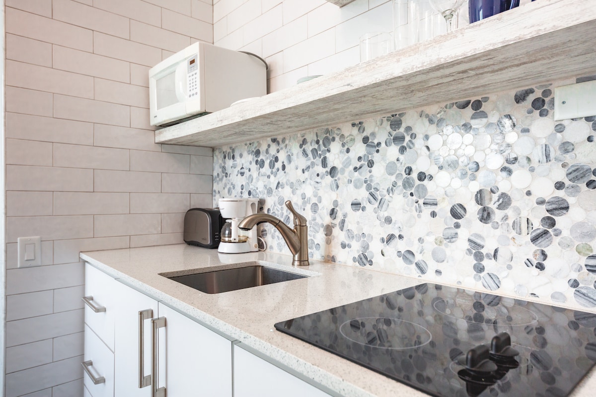 The compact kitchenette features a sleek countertop with a recessed sink and an electric cooktop for meal preparation. Above, a modern microwave is installed, and a mosaic tile backsplash adds visual interest with varying shades of blue and gray. Glassware is neatly arranged on the shelves.