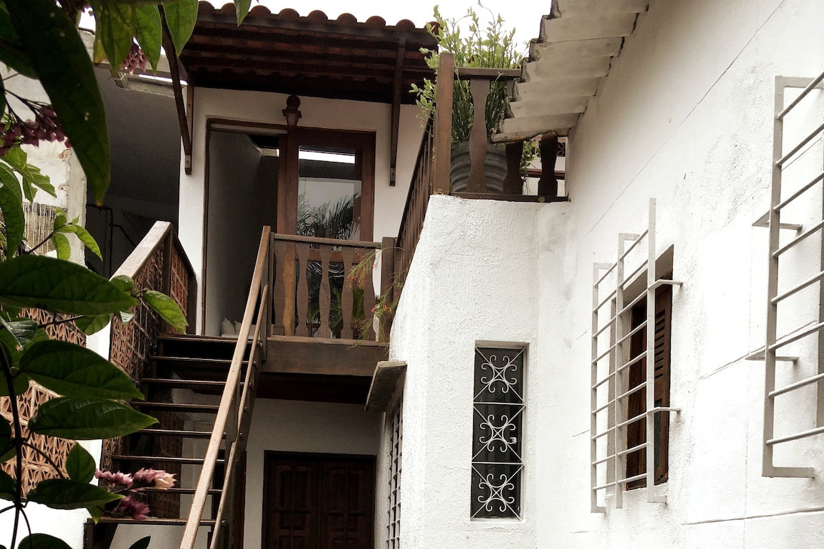 An entrance with wooden stairs leads to a second-level area, surrounded by greenery. The white exterior walls feature decorative windows, while a sheltered porch area fosters a connection with nature.