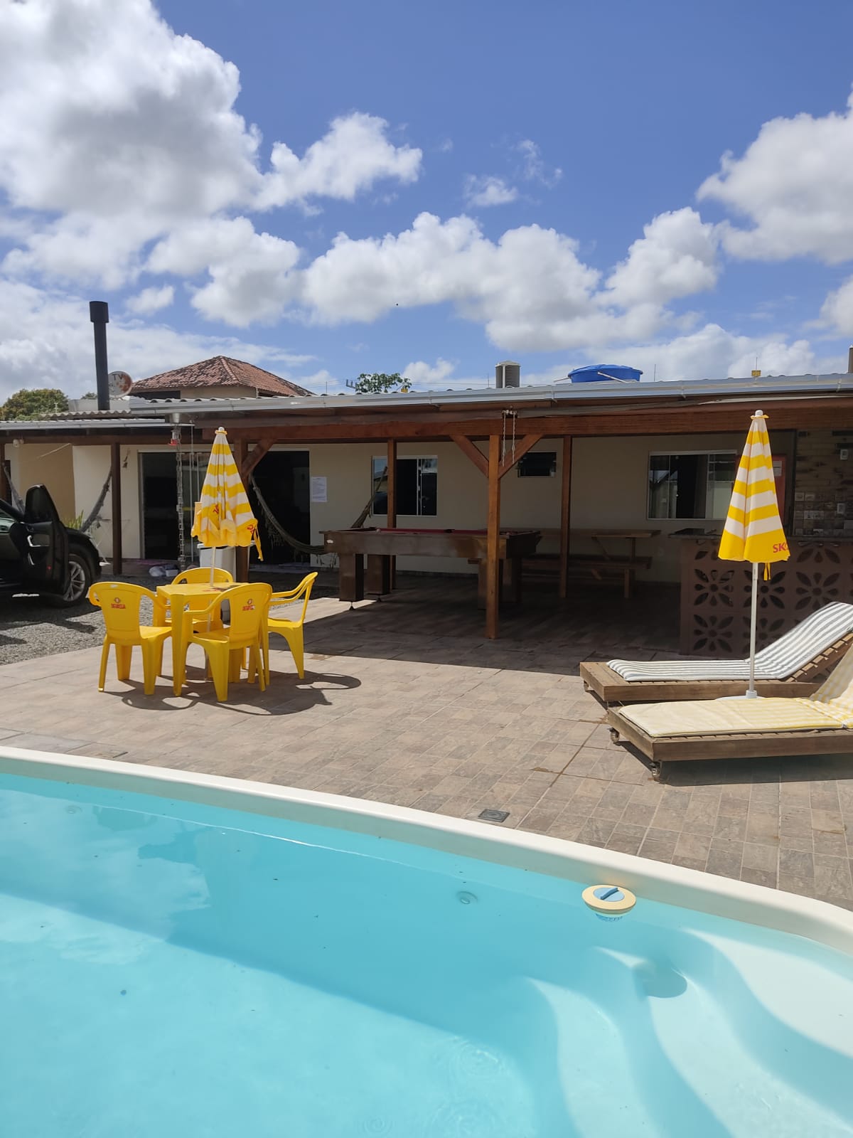 A private outdoor area features a refreshing swimming pool with a tiled deck. Yellow umbrellas provide shade over a dining set for four. A lounge chair is positioned nearby for relaxation. The building is set against a backdrop of bright blue sky with fluffy clouds.