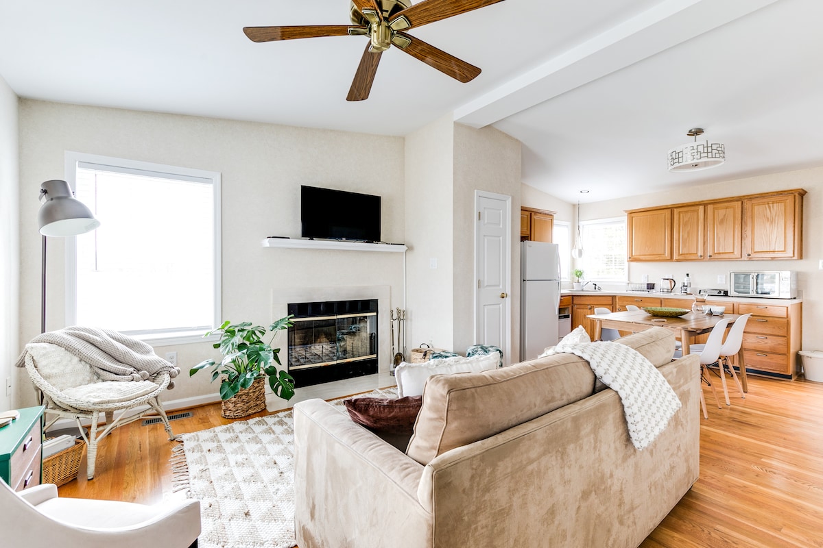 A bright living area features a comfortable sofa facing a wall-mounted television above a decorative fireplace. Light wood cabinets and countertops are visible in the kitchen area, complemented by a natural wood floor. A cozy armchair and a patterned area rug enhance the welcoming atmosphere.