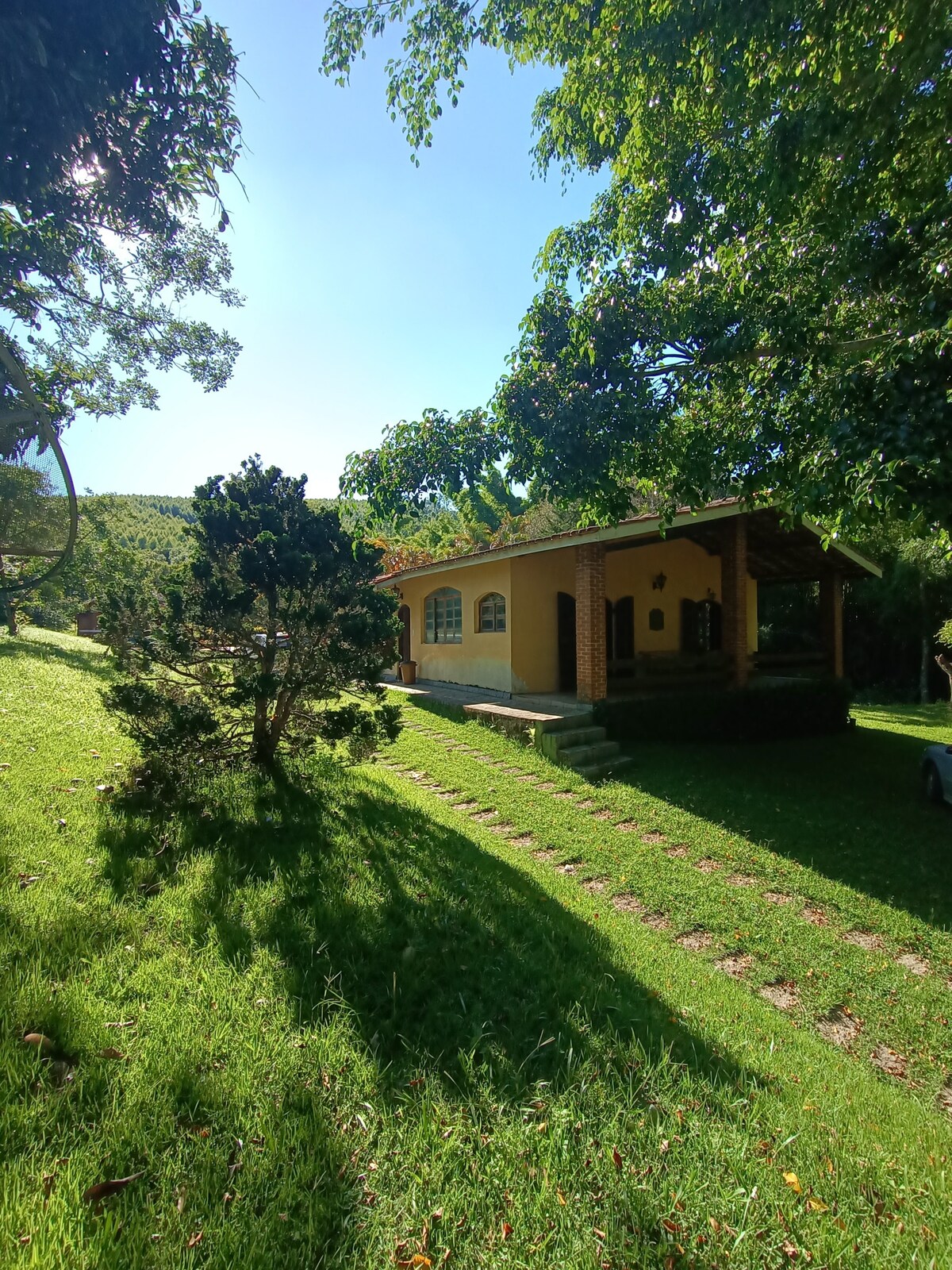 A one-story cottage is set amidst lush green grass and trees, featuring a welcoming porch and clear blue sky overhead. Sunlight filters through the leaves, casting soft shadows on the ground. A stone path leads to the entrance, enhancing accessibility in the natural surroundings.
