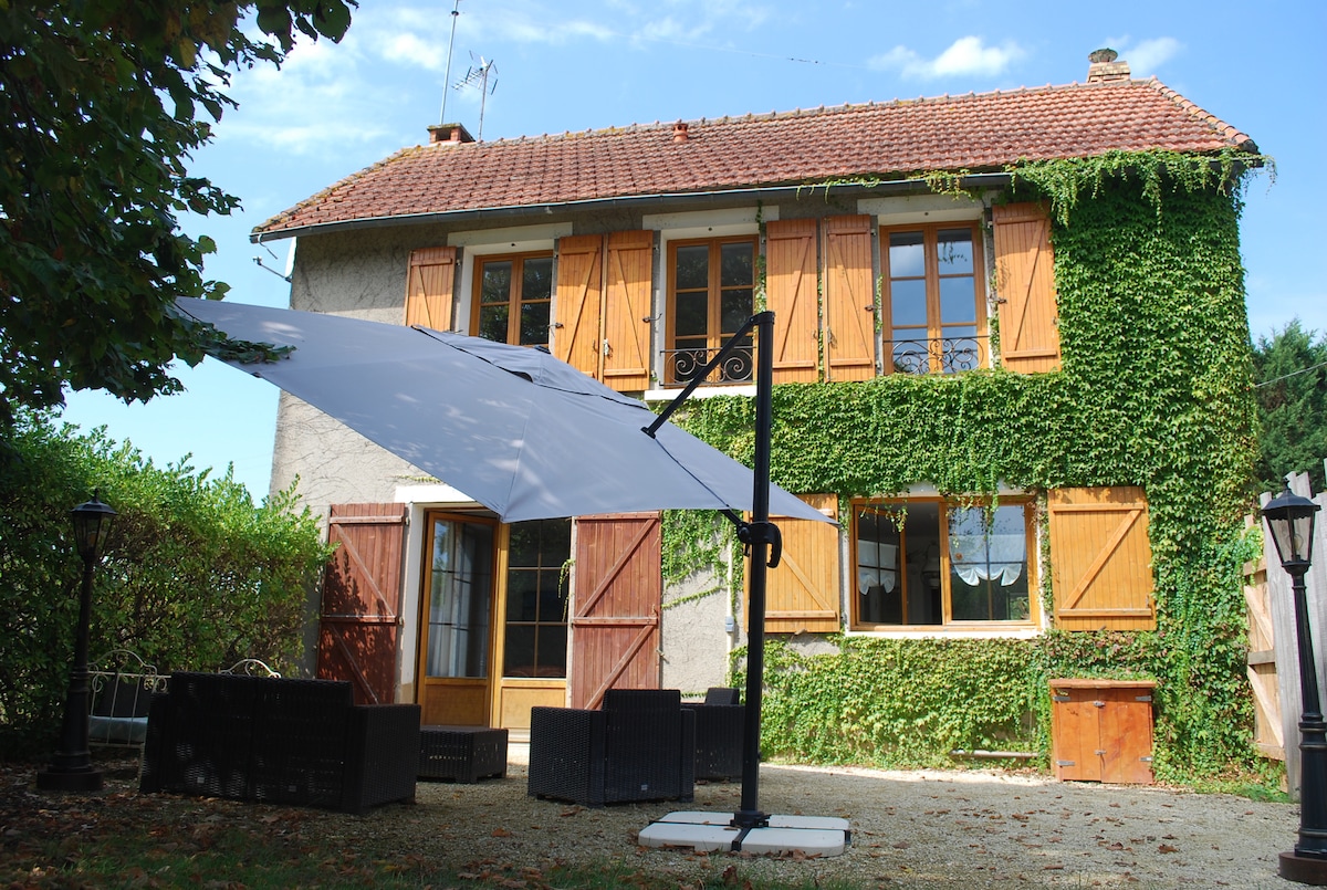 The exterior of a charming two-story farmhouse is visible, featuring ivy-covered walls and wooden shutters. A shaded seating area with modern furniture is positioned in front, complemented by a decorative lamp post. The inviting entrance is framed by double doors, creating a welcoming atmosphere.