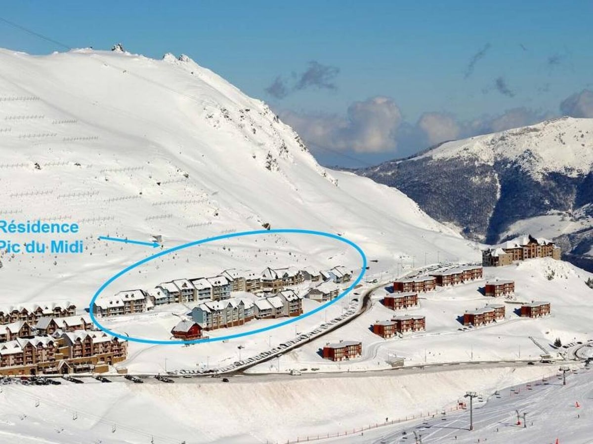 A snow-covered landscape showcases a cluster of buildings nestled in a curved formation. The architecture reflects a harmonious blend of wood and stone. The surrounding mountains create a serene backdrop, illustrating a peaceful winter environment.
