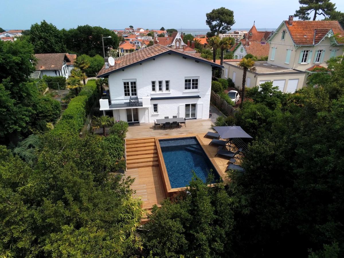 An aerial view of a renovated house surrounded by lush greenery and trees, featuring a deck and a swimming pool. The pool is framed by a wooden terrace, and outdoor seating is visible, promoting a serene and private outdoor setting.