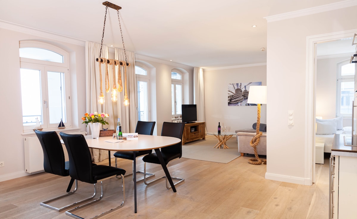 An open-plan living and dining area is depicted, featuring a round dining table surrounded by four black chairs. A comfortable sofa is positioned across from a television, with light flooding in through large windows. The neutral decor is complemented by decorative elements on the table.