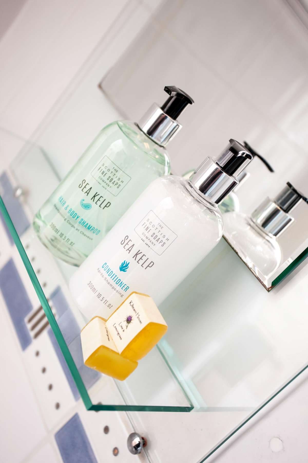 A mirrored shelf displays two bottles of sea kelp shampoo and conditioner, designed with clear labels. Beside them, two small bars of soap are neatly placed, emphasizing the functional and aesthetic arrangement of toiletries.