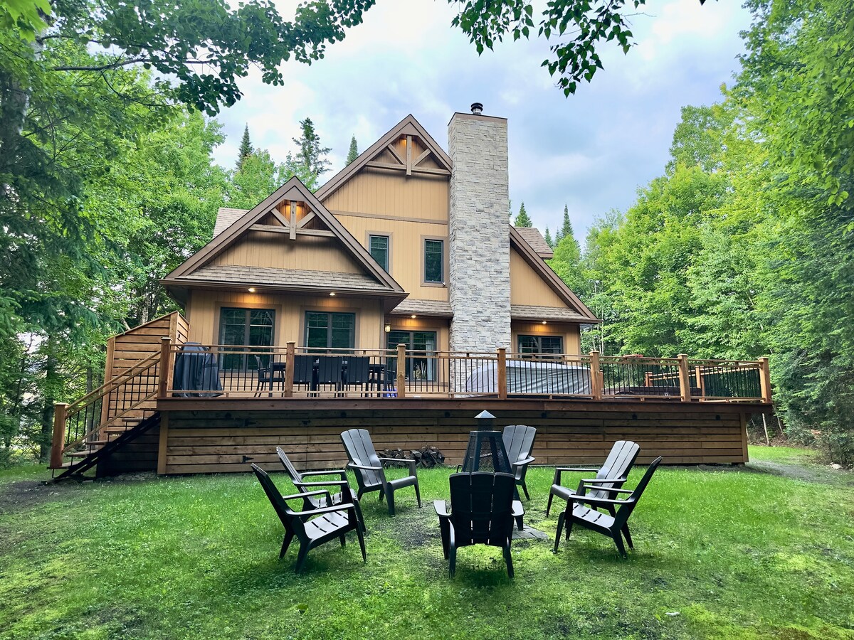 A modern chalet is positioned among lush green trees, featuring a spacious wooden deck with a stone chimney. A fire pit area with several black Adirondack chairs is situated in the foreground, providing a cozy gathering space for outdoor relaxation.