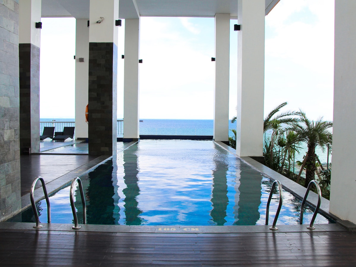 An outdoor pool is framed by sleek stone walls, reflecting the serene blue of the ocean beyond. Lounge chairs are positioned nearby, and palm trees sway gently in the background, enhancing the coastal view. Natural light fills the space, creating a bright atmosphere.