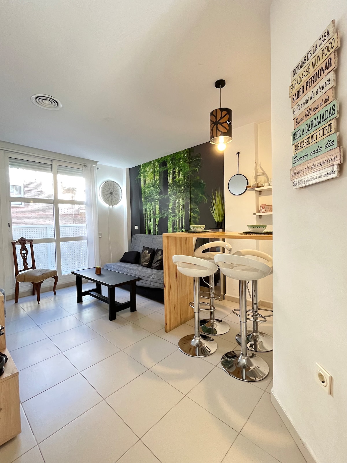 A modern living area features a spacious gray sofa against a wall adorned with a vibrant forest mural. A wooden bar table with two high stools is positioned next to it, with natural light illuminating the space through large windows.