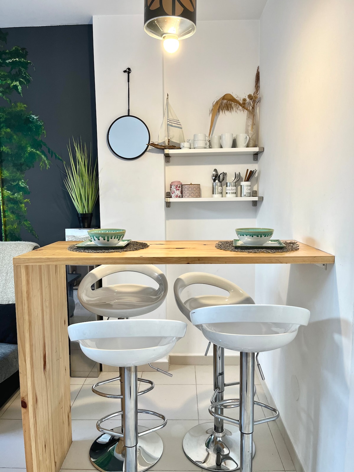 A wooden bar table is paired with three modern, chrome bar stools. The wall shelf above is adorned with various tableware and decor items, including a small round mirror, creating a functional yet stylish dining area.
