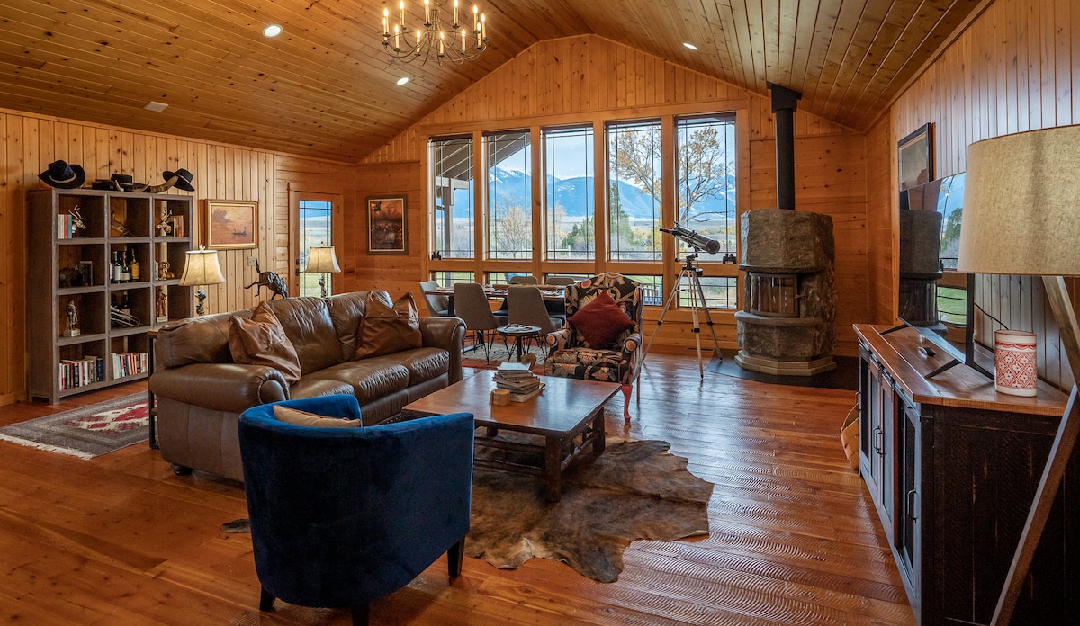 A warmly styled living room features high vaulted wood ceilings and large windows offering expansive views of the surrounding mountains. A comfortable leather sofa and armchair are positioned around a wooden coffee table, complemented by a decorative rug. A wood stove adds rustic charm.
