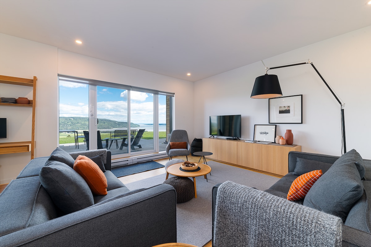 A modern living area features a comfortable gray seating arrangement paired with decorative orange pillows. A wooden media console displays a television. Large sliding glass doors invite natural light and provide views of the outdoor space, complemented by a stylish floor lamp.