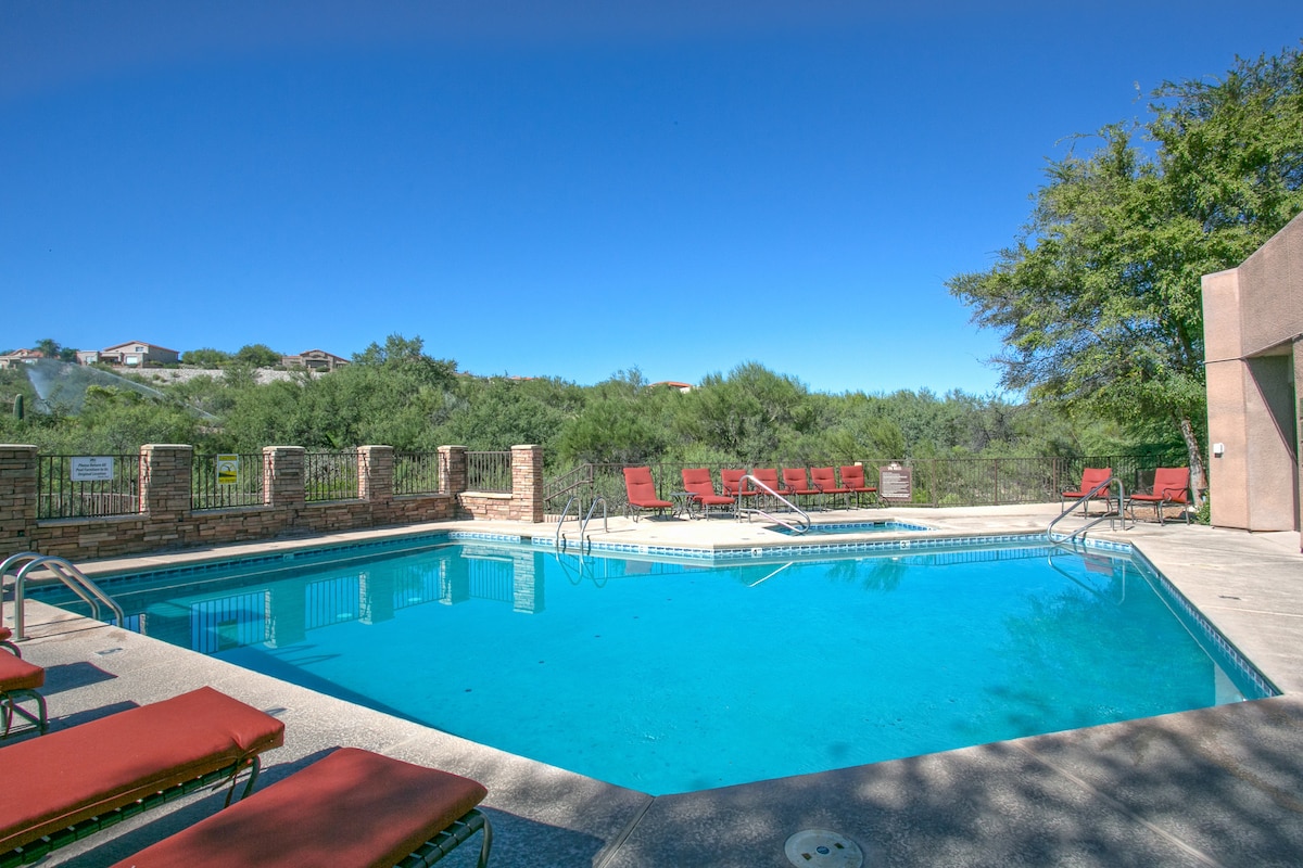 A community pool is surrounded by comfortable lounge chairs, inviting relaxation. Lush greenery and distant hills create a serene backdrop against a clear blue sky. The pool area is well-maintained, showcasing a spacious environment for outdoor enjoyment.