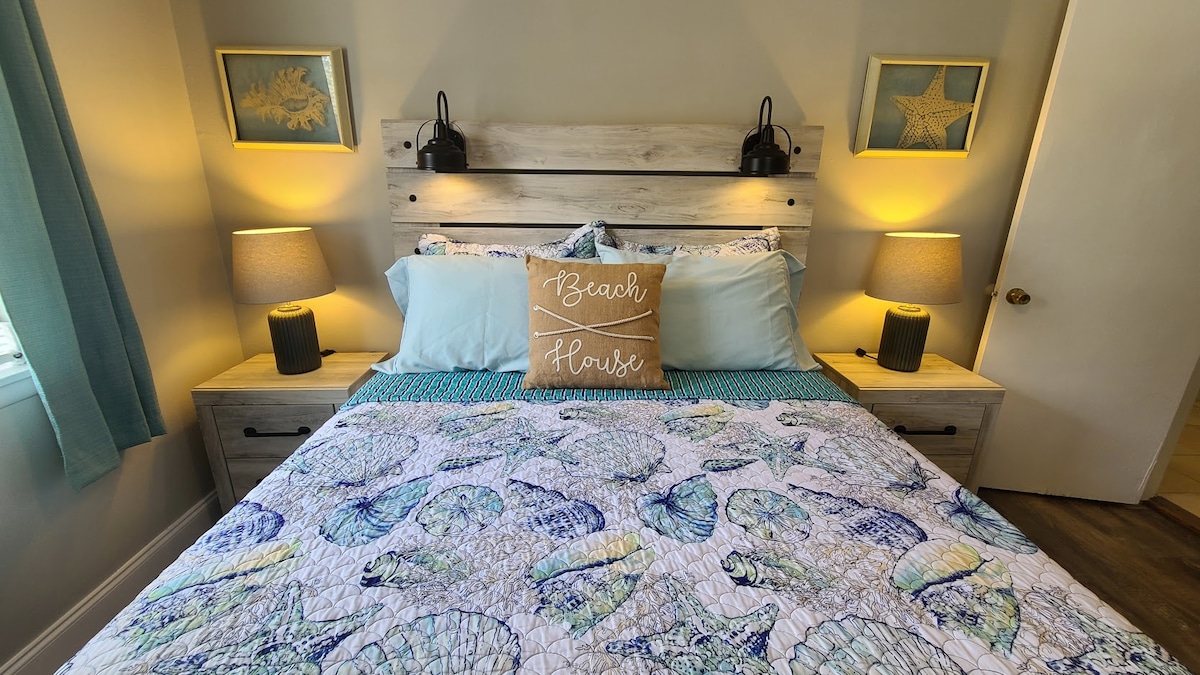 A cozy bedroom features a wooden headboard with two bedside lamps. A quilted bedspread adorned with seashells covers the spacious bed, complemented by light blue pillows. Framed coastal artwork is displayed on the walls, and soft natural light filters through the window dressed with sheer curtains.