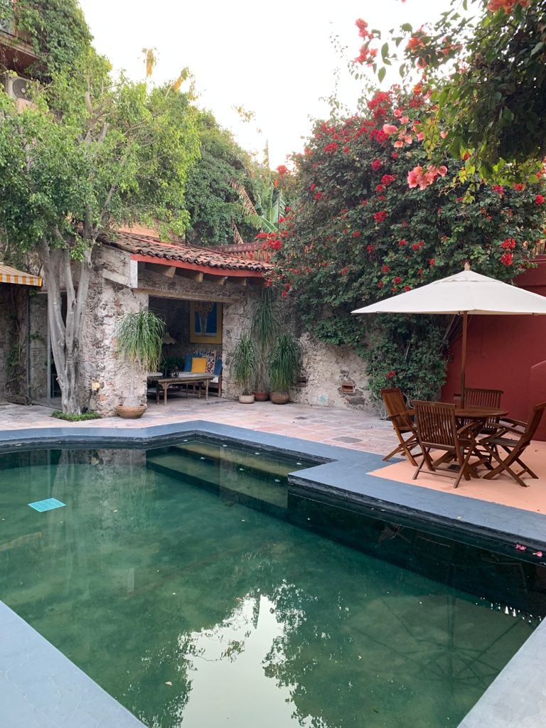 A tranquil courtyard features a functional pool surrounded by lush greenery and vibrant bougainvillea. Outdoor dining furniture is arranged nearby under a large umbrella, inviting relaxation amid the serene environment. The rustic stone walls provide a charming backdrop to the space.
