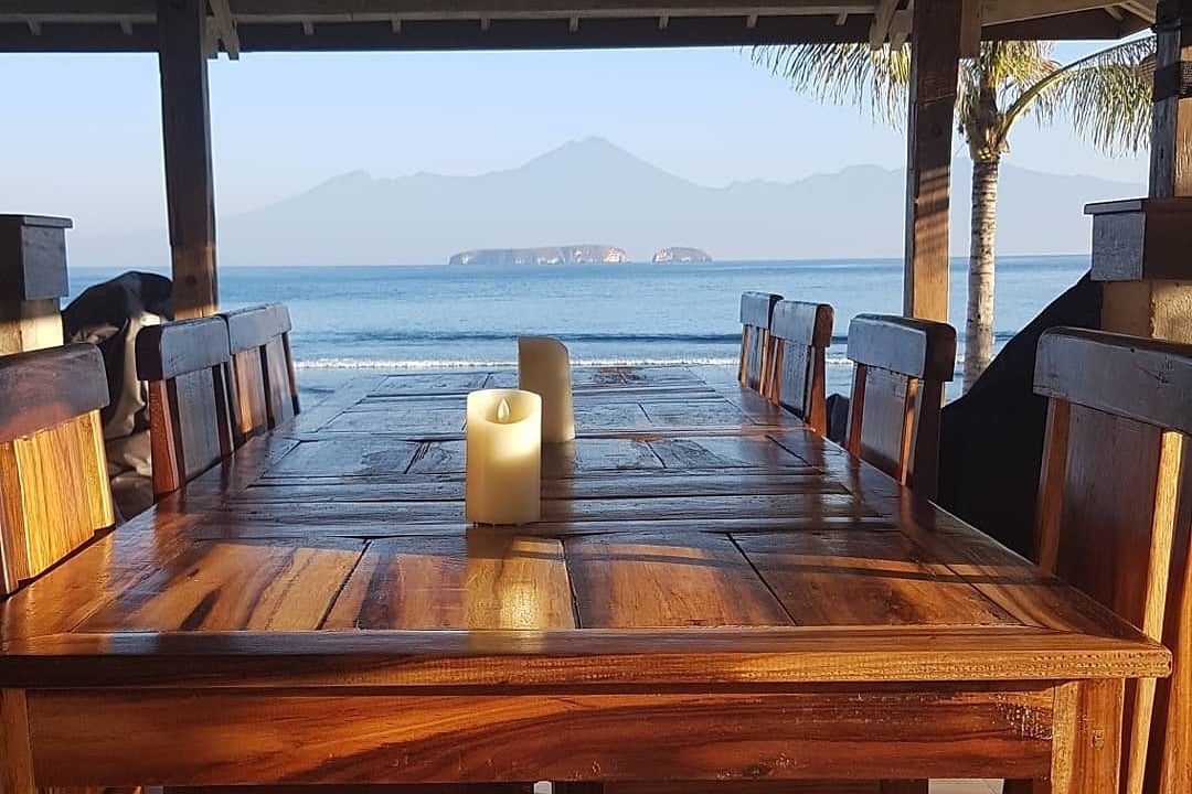 A wooden dining table is positioned under a shaded structure, overlooking the beach. A lit candle provides a gentle glow. In the distance, Pulau Dua Islands and Mt. Rinjani are visible against a calm blue sea and clear sky.