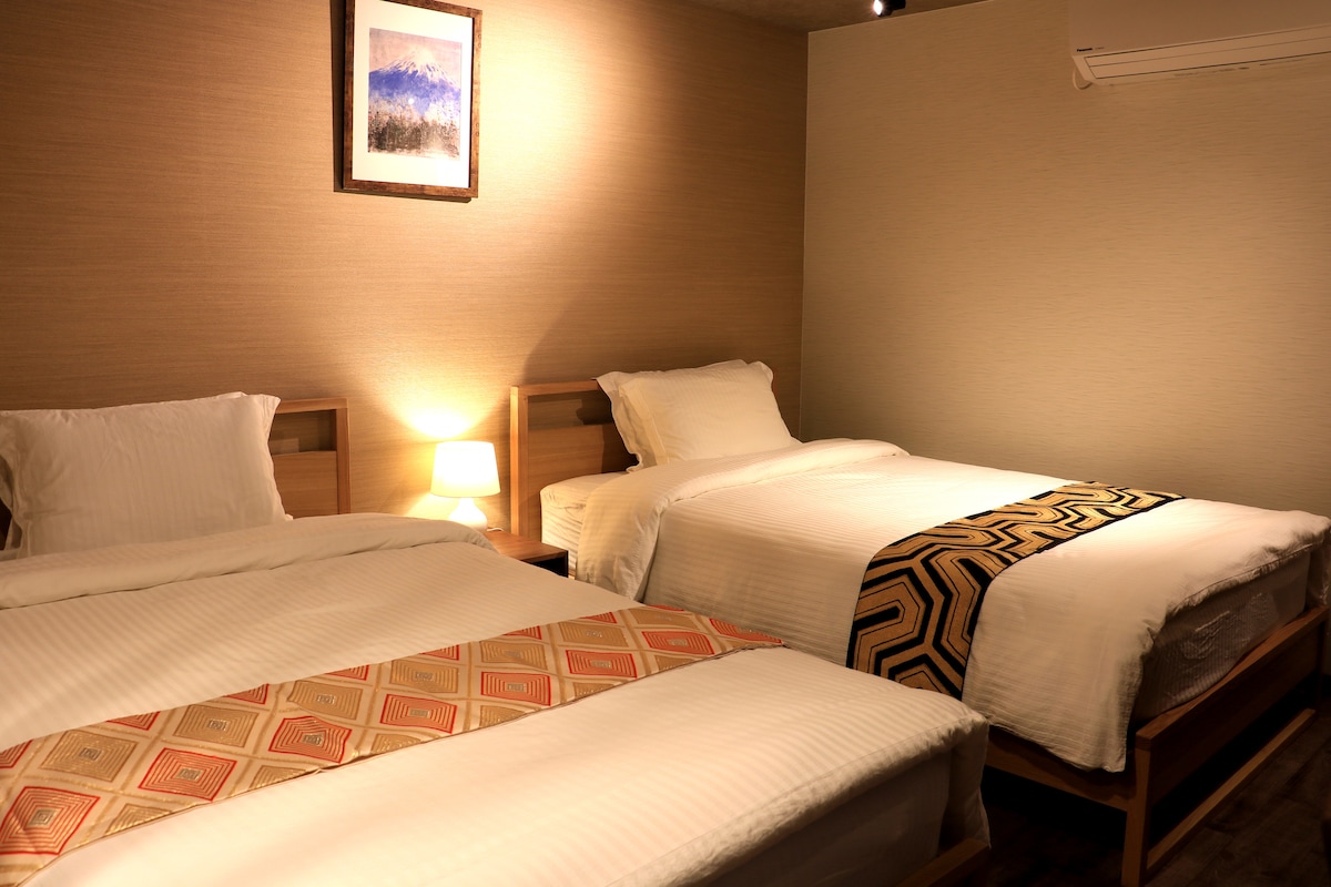 The sleeping area features two spacious beds, each dressed in crisp white linens. A subtle patterned runner decorates each bed. A bedside lamp provides soft illumination, while a framed landscape artwork adds a touch of interest to the neutral-toned wall.