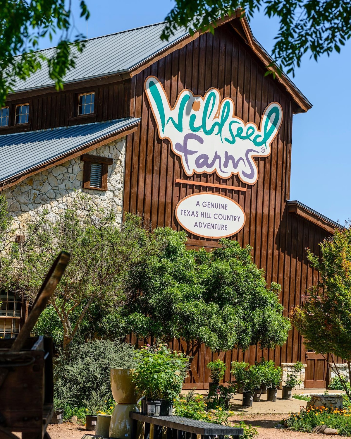 The image showcases a rustic building with a stone and wood facade, prominently featuring the Wildseed Farms sign. Lush greenery surrounds the structure, enhancing its natural charm. A clear blue sky creates a bright backdrop, complementing the inviting atmosphere of the area.