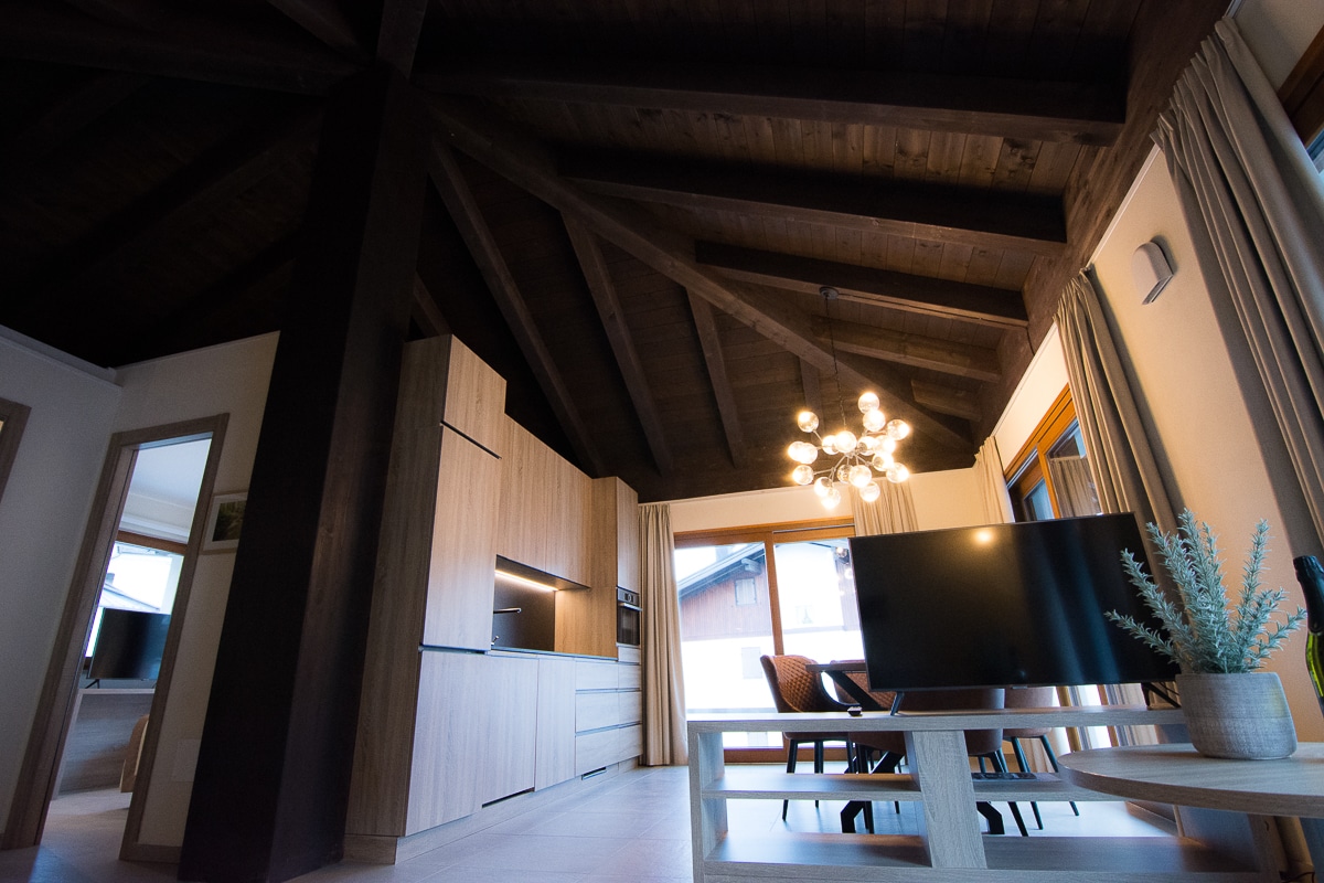 The interior features a high, wooden ceiling with exposed beams, creating a spacious feeling. A modern TV sits on a light wooden table, while two woven chairs are positioned nearby. Natural light filters through large windows adorned with simple curtains, illuminating the room.