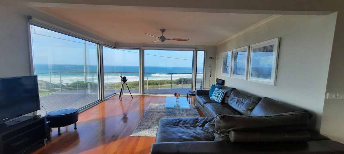 A spacious living area is designed with large glass windows, offering unobstructed views of the ocean and coastline. A comfortable sectional sofa is positioned around a coffee table, and a television sits on a stand nearby. A decorative rug adds warmth to the wooden floor.