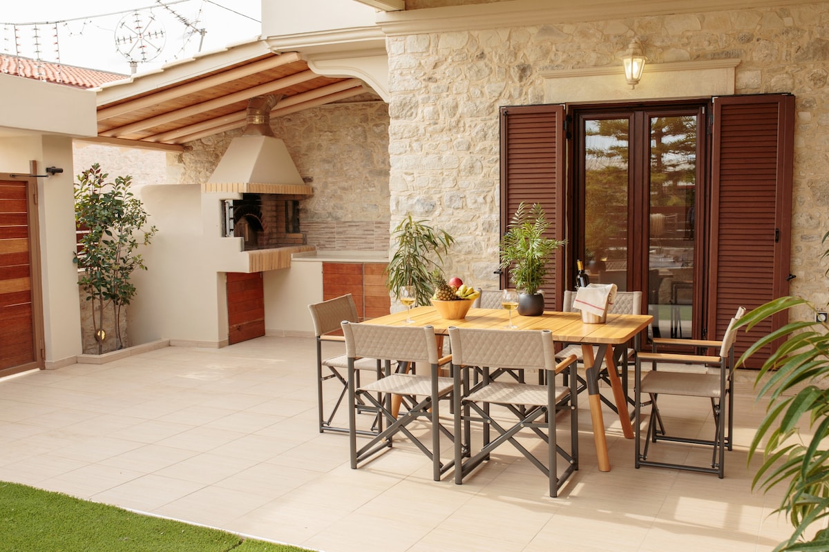 A spacious outdoor terrace features a wooden dining table surrounded by comfortable chairs. A barbeque area is positioned nearby, with stone details enhancing the space. Lush green plants are placed around, contributing to a relaxed and inviting atmosphere.