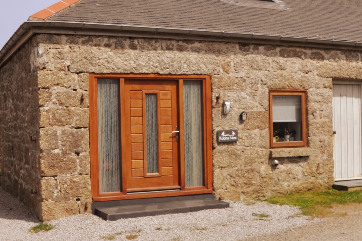 The entrance to Robins Nest features a wooden door with large vertical panels, complementing the rustic stone façade. A small window beside the door reveals a glimpse of greenery inside. The entrance is accessed via a short path lined with gravel.