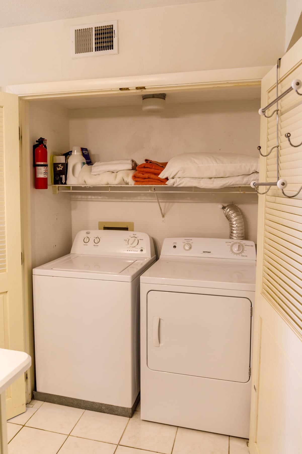 A compact laundry area features a stacked washer and dryer, neatly positioned beside a shelf containing extra linens. A fire extinguisher and two additional items are visible on the shelf, contributing to the functionality of the space.