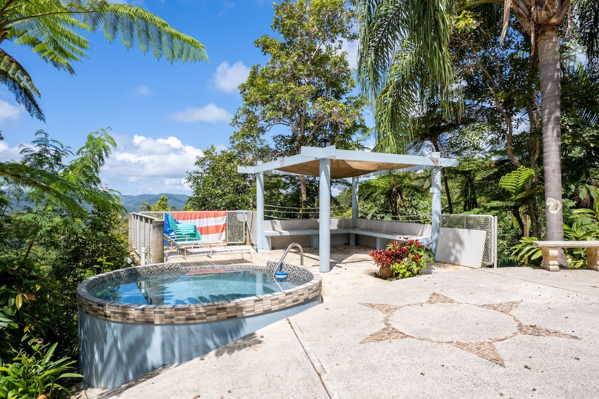 An outdoor patio features a heated jacuzzi surrounded by lush greenery, providing a serene setting. A covered lounge area is adjacent, with comfortable seating arranged under a canopy. The natural landscape and bright blue sky create a calming atmosphere.
