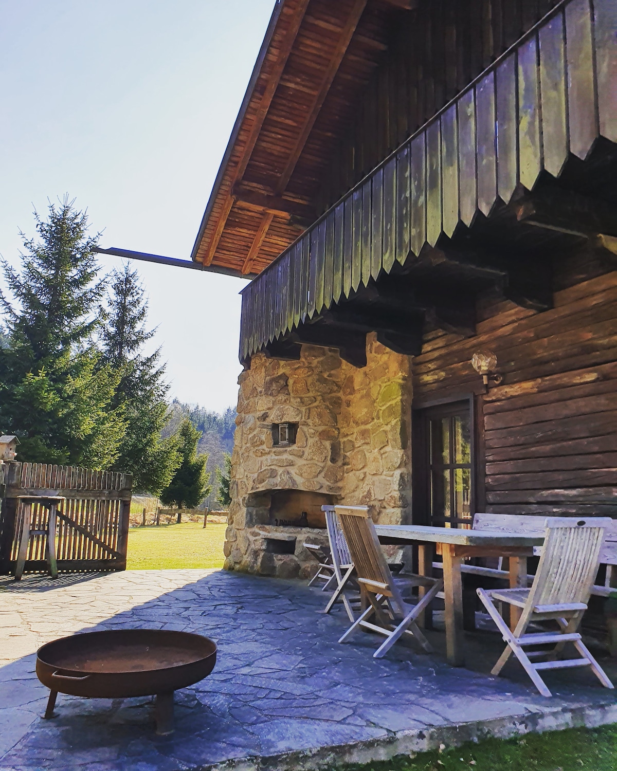 A rustic stone terrace features a wooden dining table with four matching chairs, accompanied by a fire pit on a stone surface. The wooden structure overhead is adorned with dark shingles, while surrounding trees provide a natural backdrop, enhancing the outdoor space.