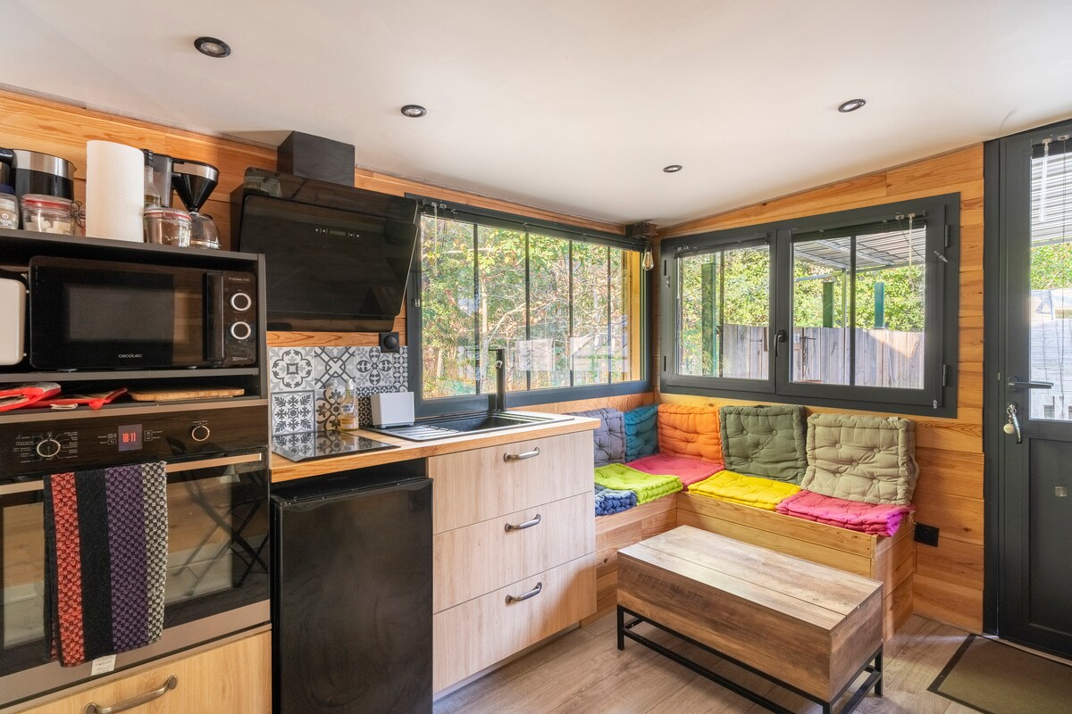 A modern kitchen area features wooden cabinetry and a black countertop. A comfortable seating nook, with colorful cushions, provides a cozy space. Natural light enters through large windows, highlighting the patterned tiles behind the stovetop. Essential appliances are neatly arranged for convenience.