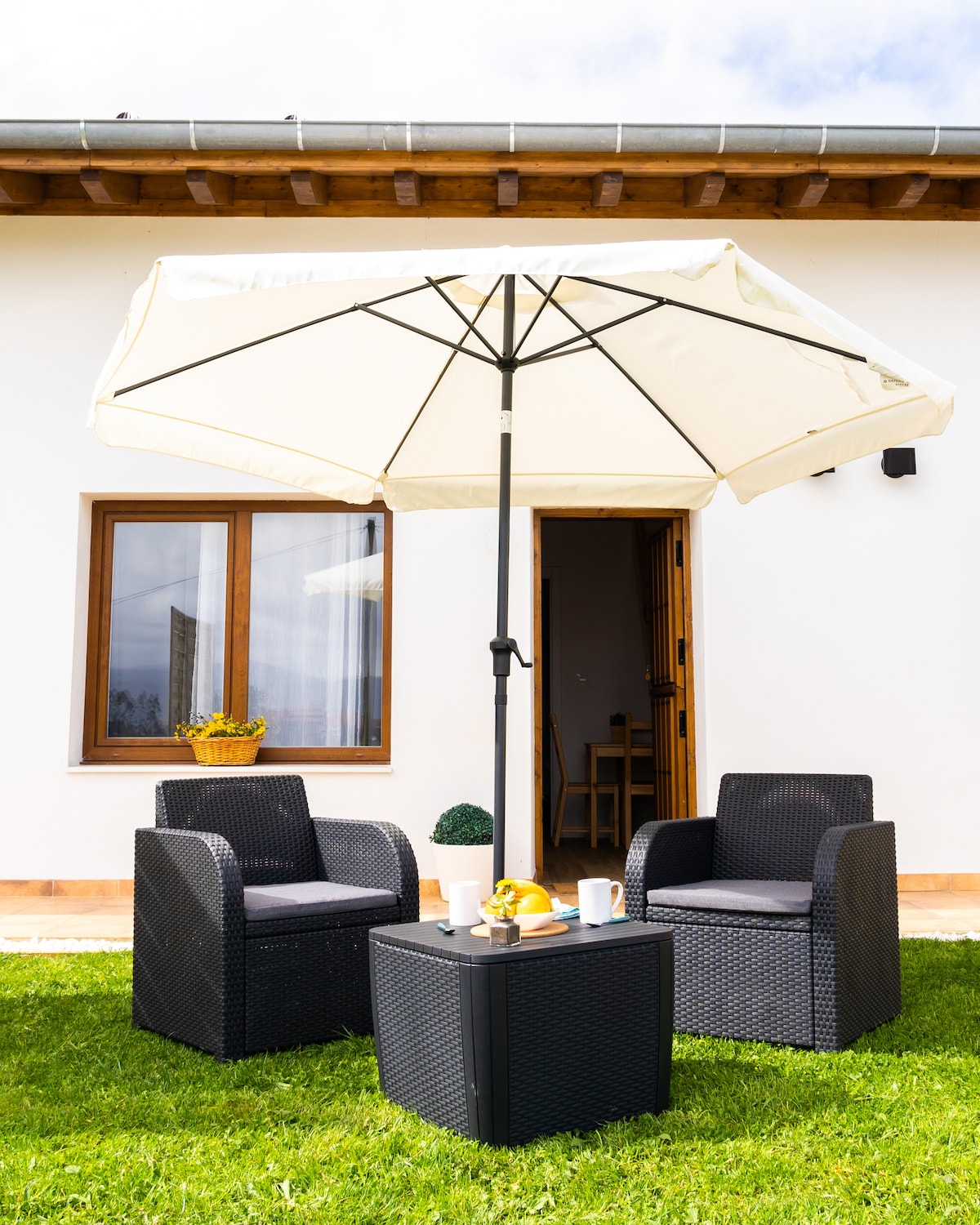 A seating area is presented in the outdoor space, featuring two dark wicker chairs and a small table. A large umbrella provides shade above the table, which holds a few cups and a plate with fruit. A cheerful flower pot is visible on a nearby window ledge.