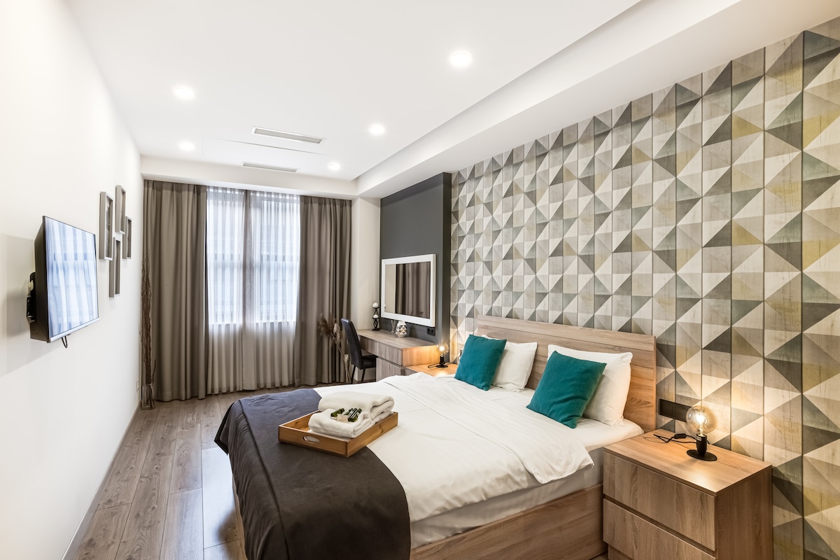 A spacious bedroom is featured, with a double bed dressed in crisp linens and accented by teal cushions. A patterned accent wall adds visual interest, while soft lighting from ceiling fixtures illuminates the room. A flat-screen TV and a desk are visible, enhancing functionality.