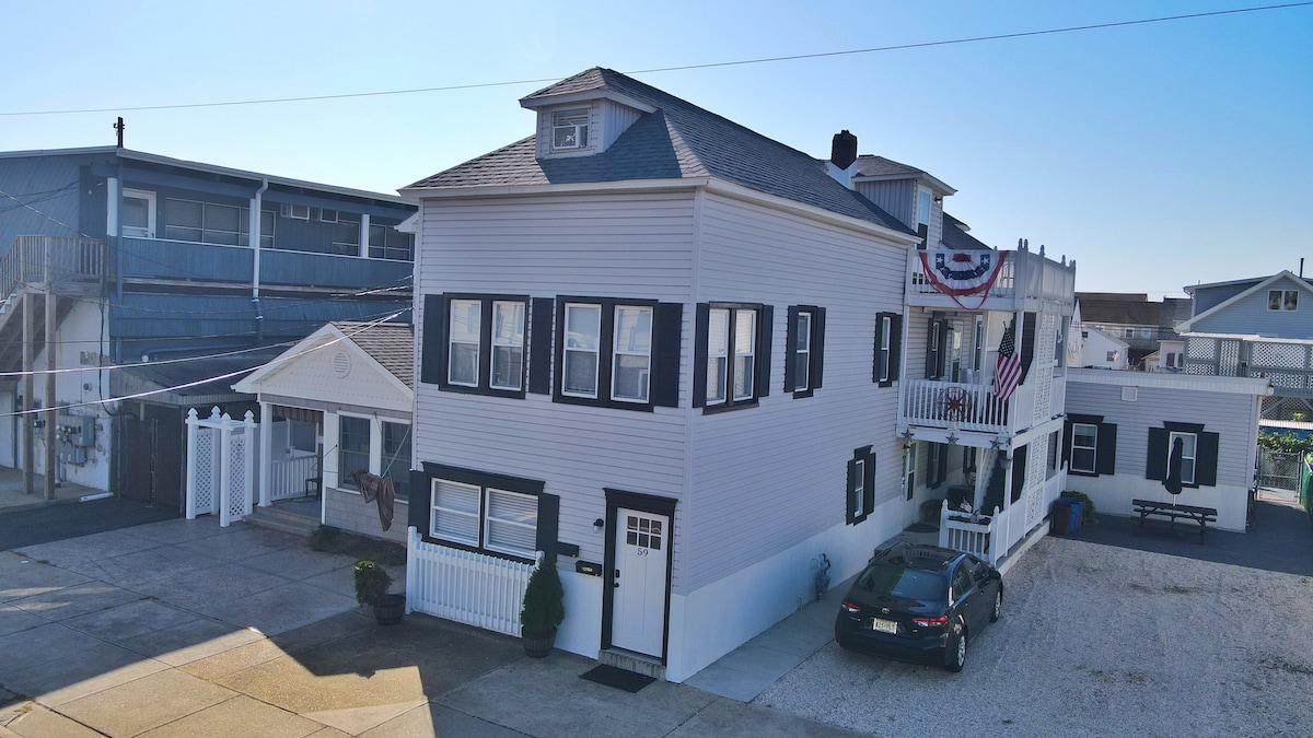 A two-story beach house is presented, featuring a light-colored exterior with black shutters. The front porch is adorned with American flags, and a gravel parking area is visible. Nearby, additional structures provide context to the neighborhood's coastal charm.