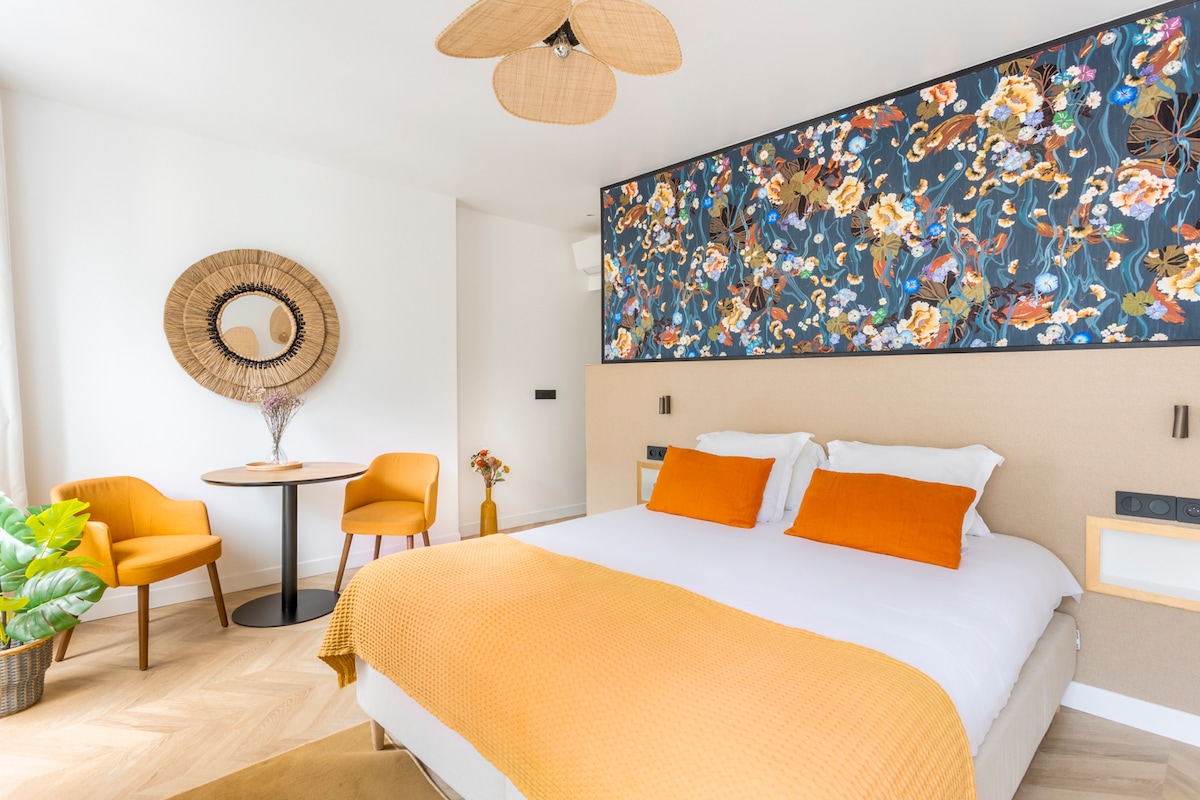 A spacious bedroom with a large bed featuring an orange blanket and accent pillows. A vibrant floral wall art piece is displayed above the bed. A round dining table with two chairs is positioned nearby, and a decorative mirror is mounted on the wall. A green plant adds a natural touch.