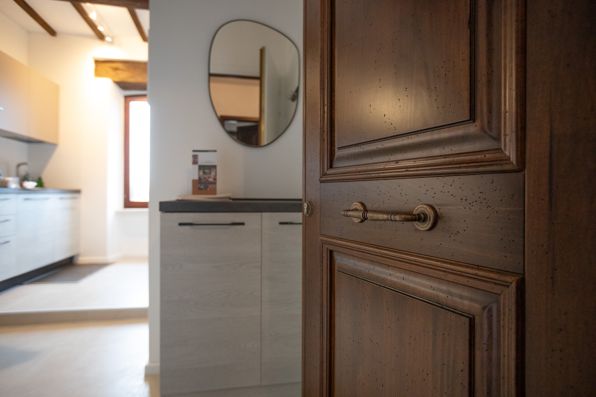 A wooden door with a brass handle is shown, leading into a modern kitchen area. The kitchen features light-colored cabinetry and a window allowing natural light to enter, creating a bright and inviting space.