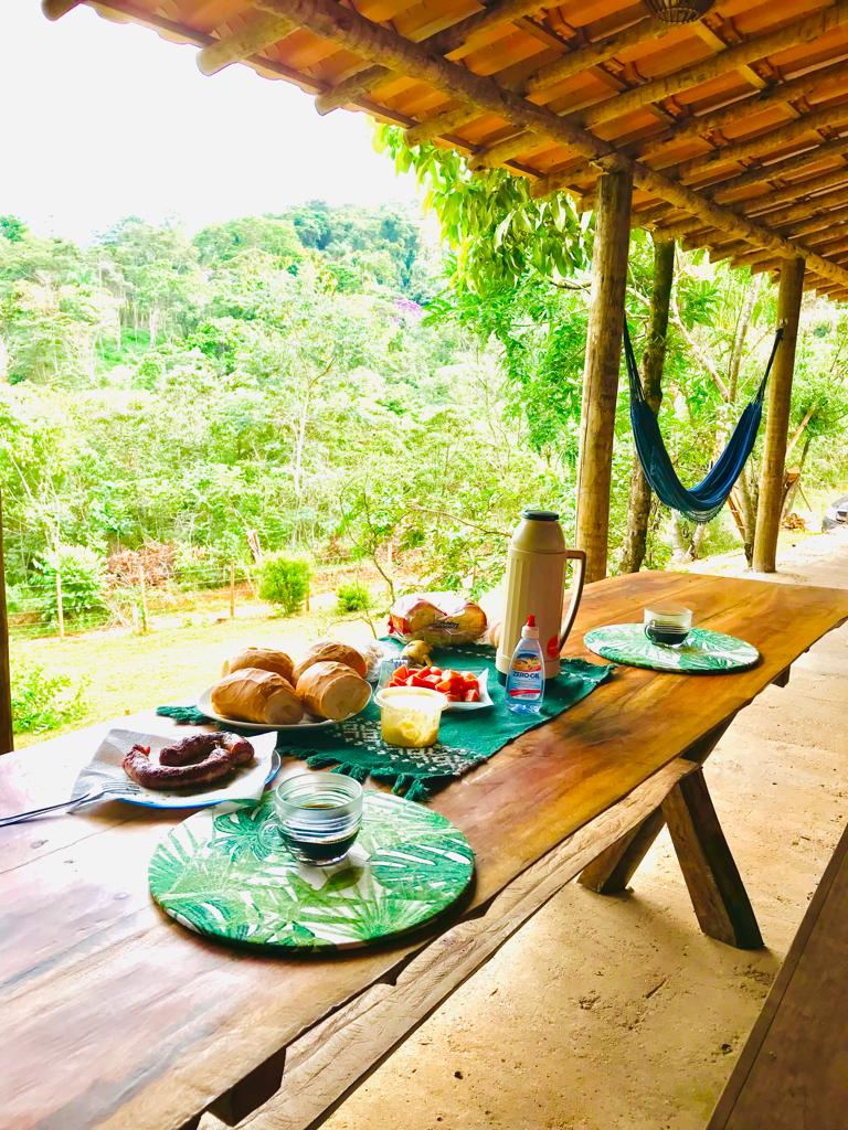 An outdoor dining area is set with a rustic wooden table adorned with green-themed placemats. A variety of breakfast items, including breads, fruit, and beverages, are displayed. Lush greenery surrounds the space, enhancing the tranquil atmosphere.