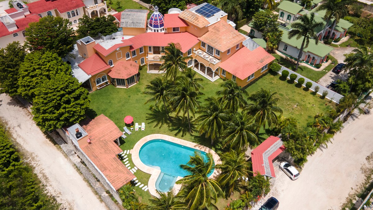 An aerial view of the property showcases a spacious layout with vibrant buildings surrounded by lush palm trees. A heart-shaped pool is centrally located, bordered by sun loungers, and well-maintained green areas provide a peaceful atmosphere.