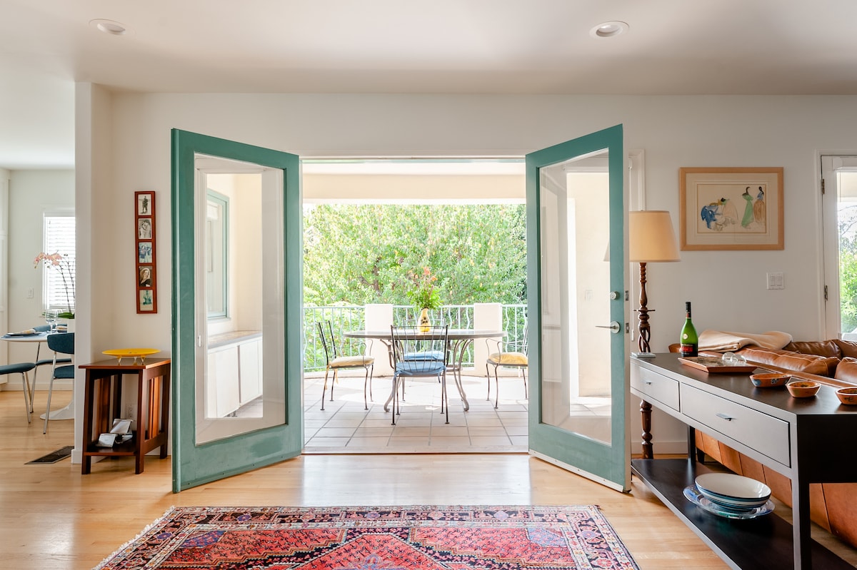 Open double doors lead to a spacious balcony overlooking greenery, with a table and chairs set for outdoor dining. Inside, a cozy living area is equipped with a light-colored sofa, a console table, and personal decor elements, resulting in a bright and inviting space.