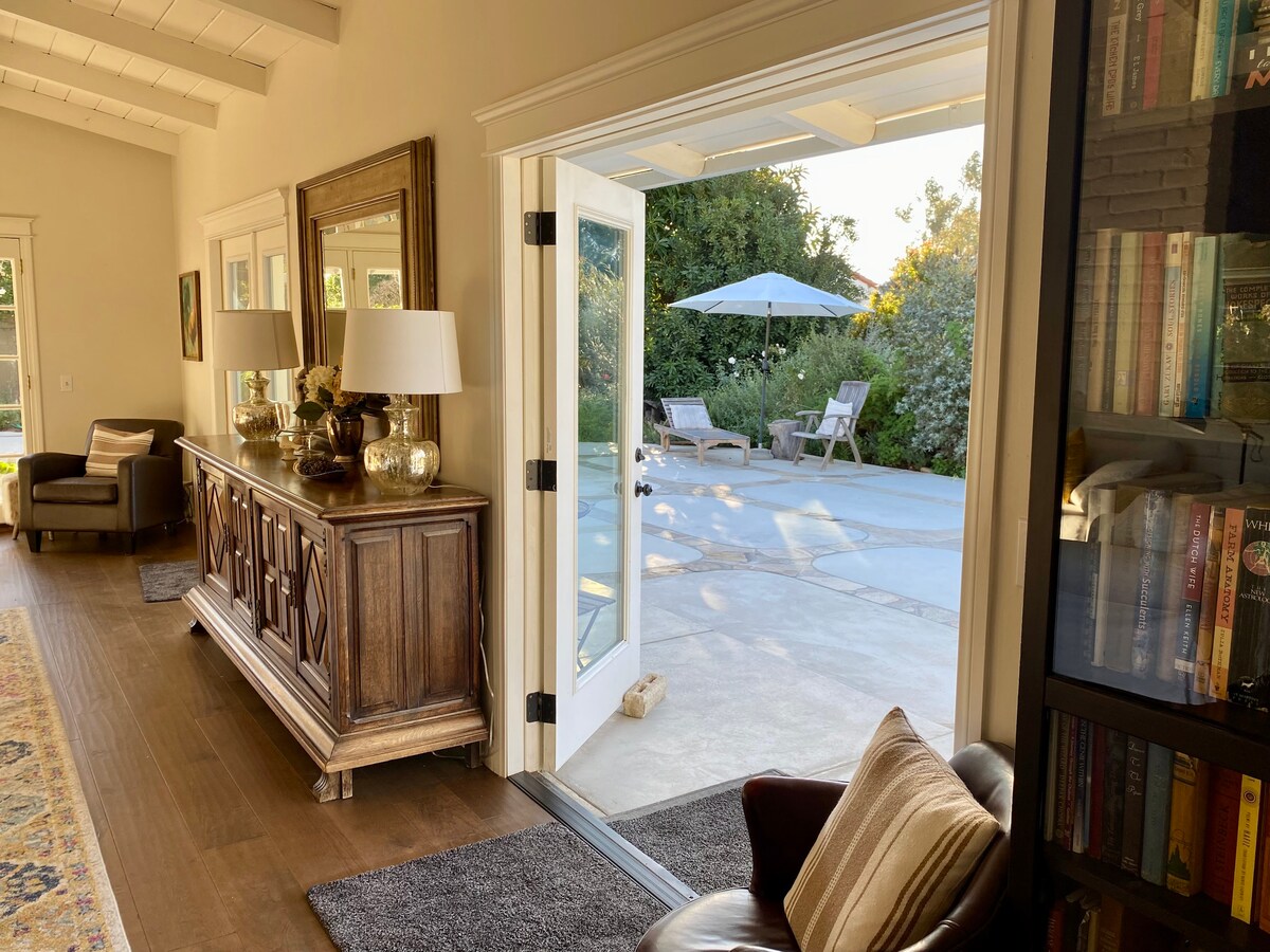 French doors open to a spacious patio area, featuring outdoor seating and an umbrella for shade. A decorative sideboard with lamps and artwork creates an inviting entryway, while natural light enhances the warm, neutral tones of the interior.